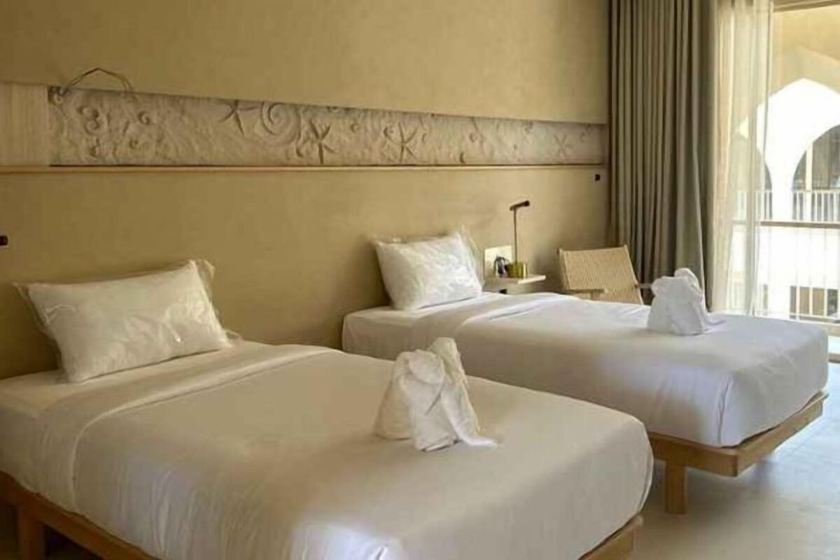 Two single beds with white linens and towels, a headboard with a decorative pattern, and a window with curtains.