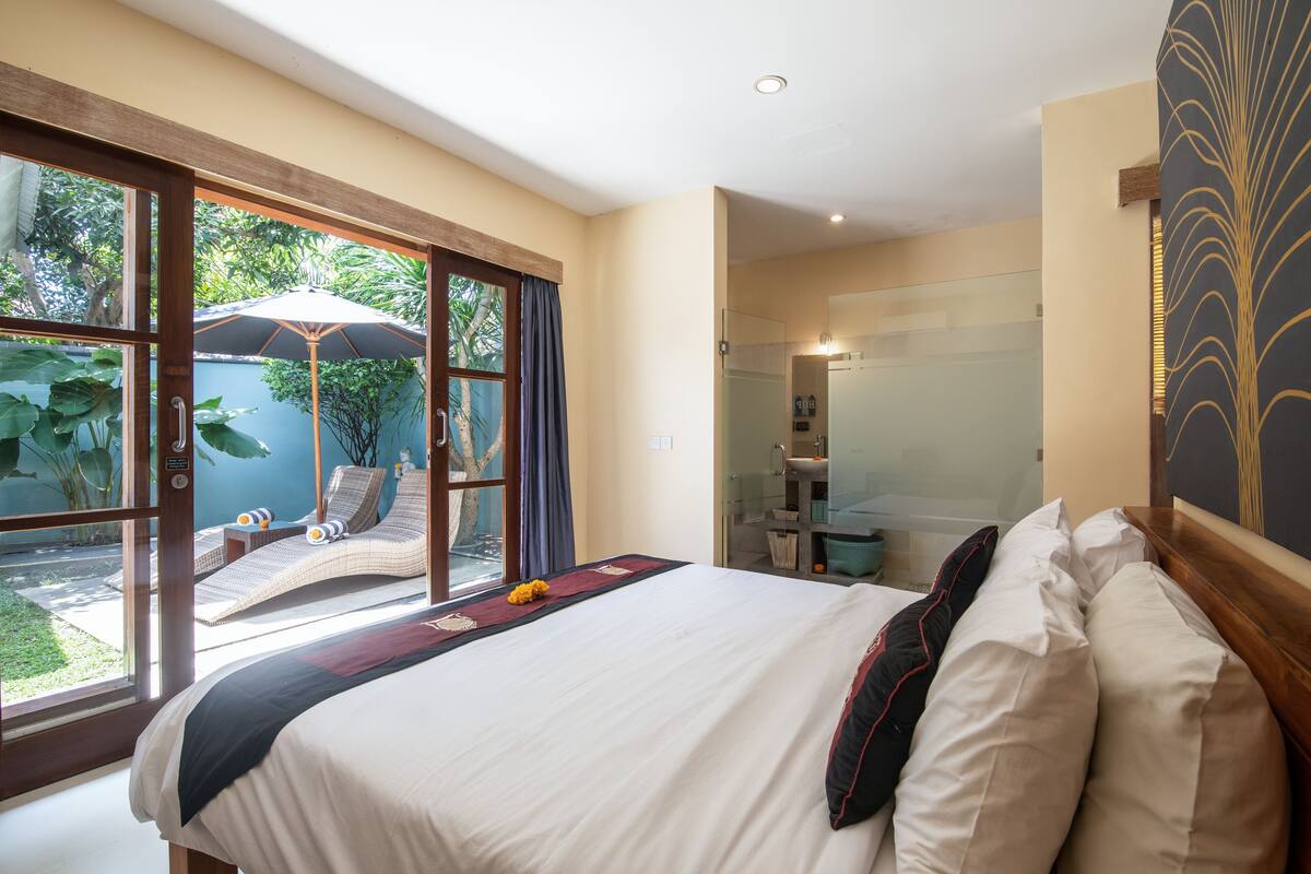 A bedroom with a large bed, a glass shower, and a view of a garden through the window.