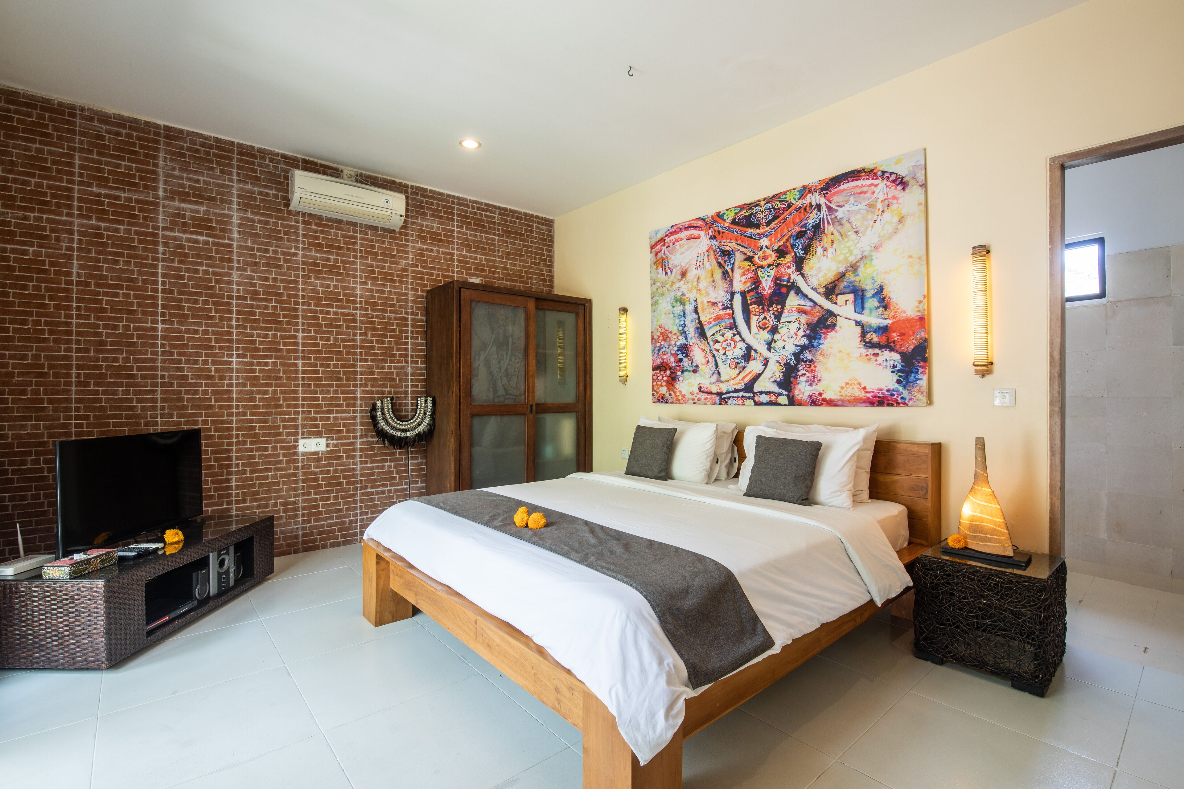 Un dormitorio con pared de ladrillo, una cama con cabecera de madera, un televisor sobre un mueble bajo y un cuadro sobre la cama.