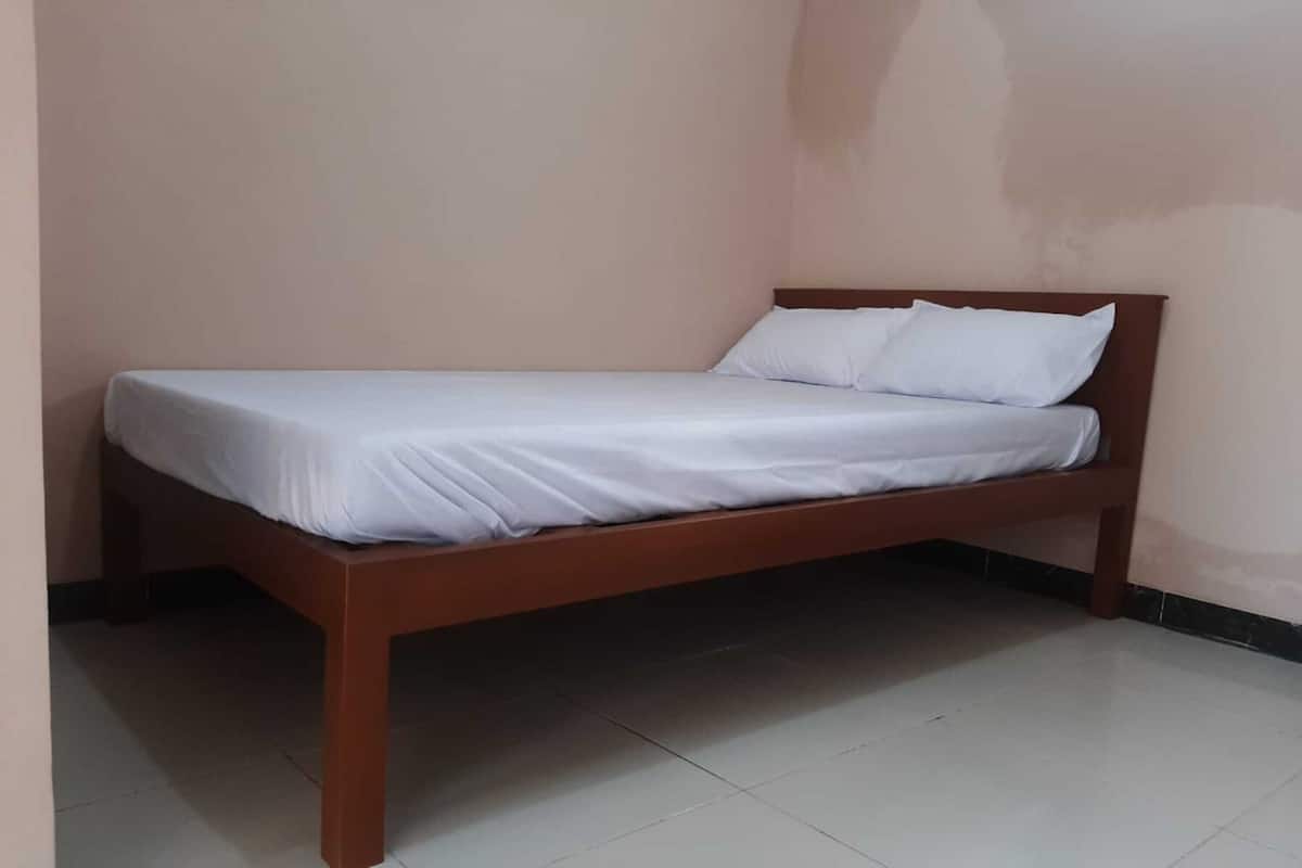 Ein Einzelbett mit Holzrahmen und weißer Bettwäsche in einem Zimmer mit schlichten Wänden und Fliesenboden.