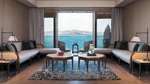 A modern hotel room with a large window offering a view of the sea, a sofa set, two side tables, and a small table with a fruit bowl. - Princes’ Palace Resort - Member of The Leading Hotels of the World (Adalar)