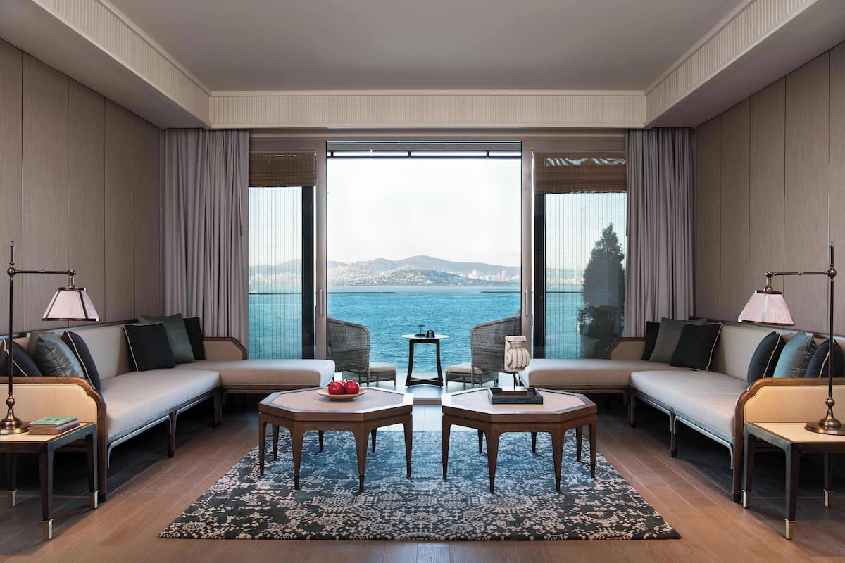 A modern hotel room with a large window offering a view of the sea, a sofa set, two side tables, and a small table with a fruit bowl.