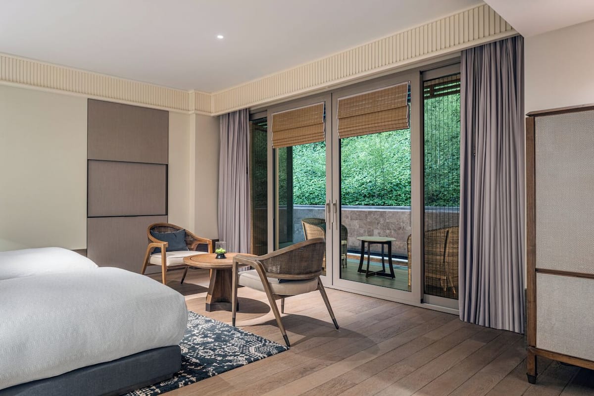 A hotel room with a bed, a desk, two chairs, a lamp, and a sliding glass door leading to an outdoor area.