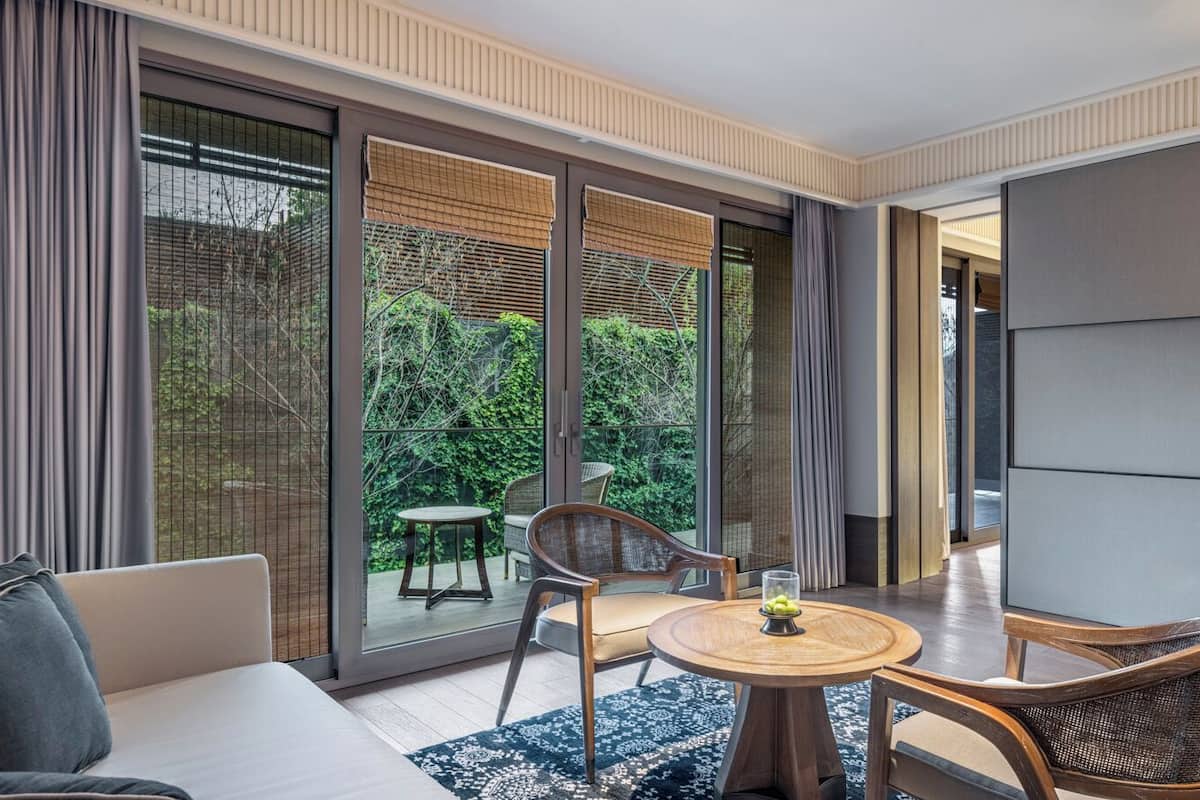 A modern hotel room with a sofa, a round table, and a chair. There is a balcony with a view of greenery.