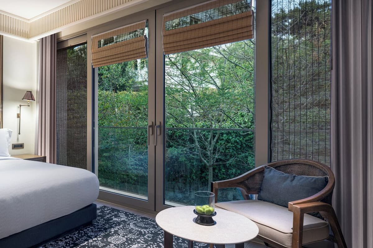 A bedroom with a bed, bedside lamps, a small round table with a fruit bowl, a chair, and a large sliding glass door leading to a balcony with greenery.