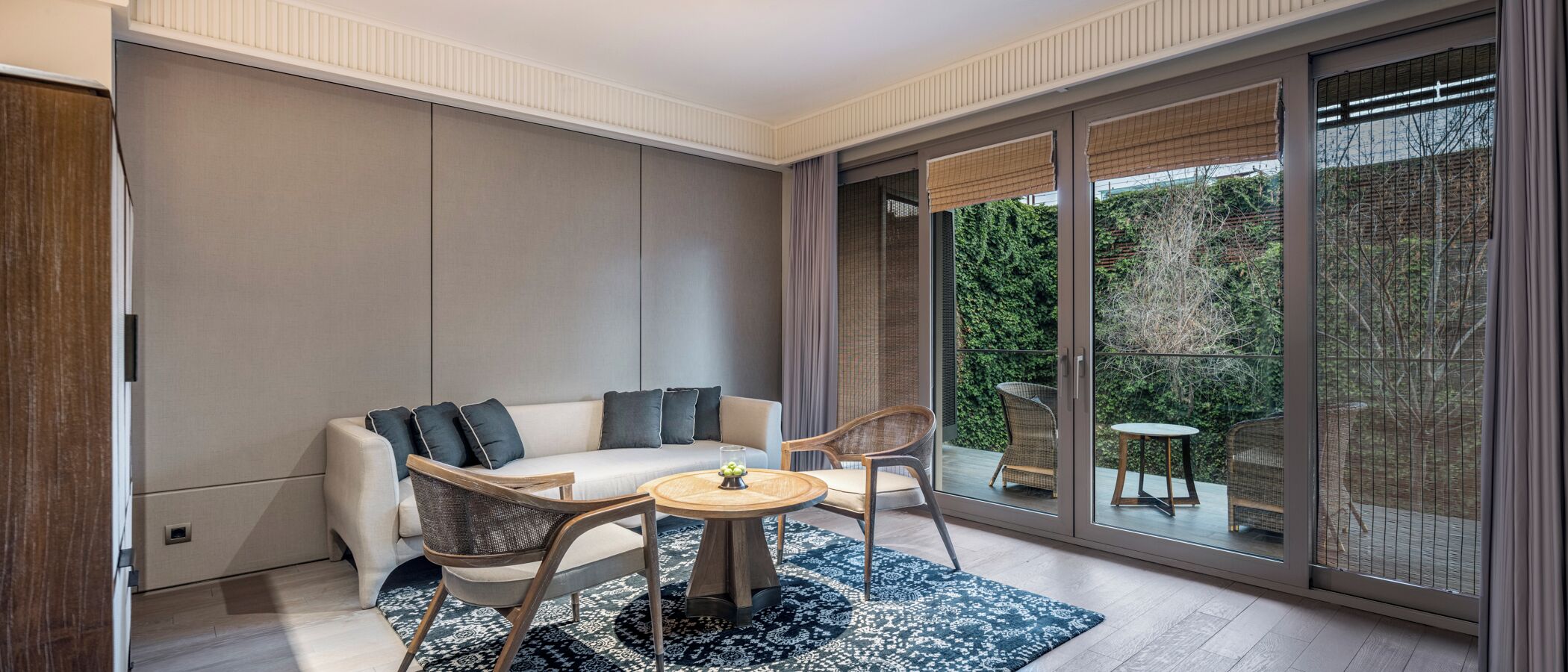 Una sala de estar moderna con un sofá, dos sillones, una mesa redonda y un amplio ventanal corredero que da a un espacio exterior con vistas a la vegetación.