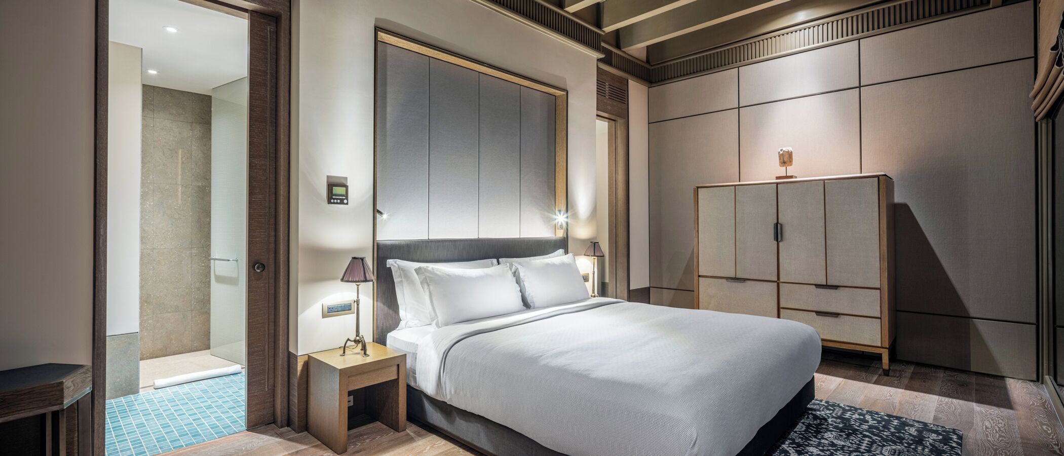 A modern hotel room with a large bed, a bedside table with a lamp, a wardrobe, and a shower area.