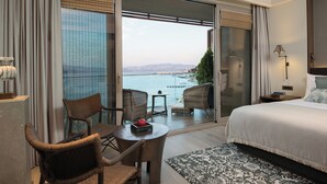 A hotel room with a large bed, a dining table with chairs, a small table with a red bowl, and a view of the sea through a sliding glass door. - Princes’ Palace Resort - Member of The Leading Hotels of the World (Adalar)