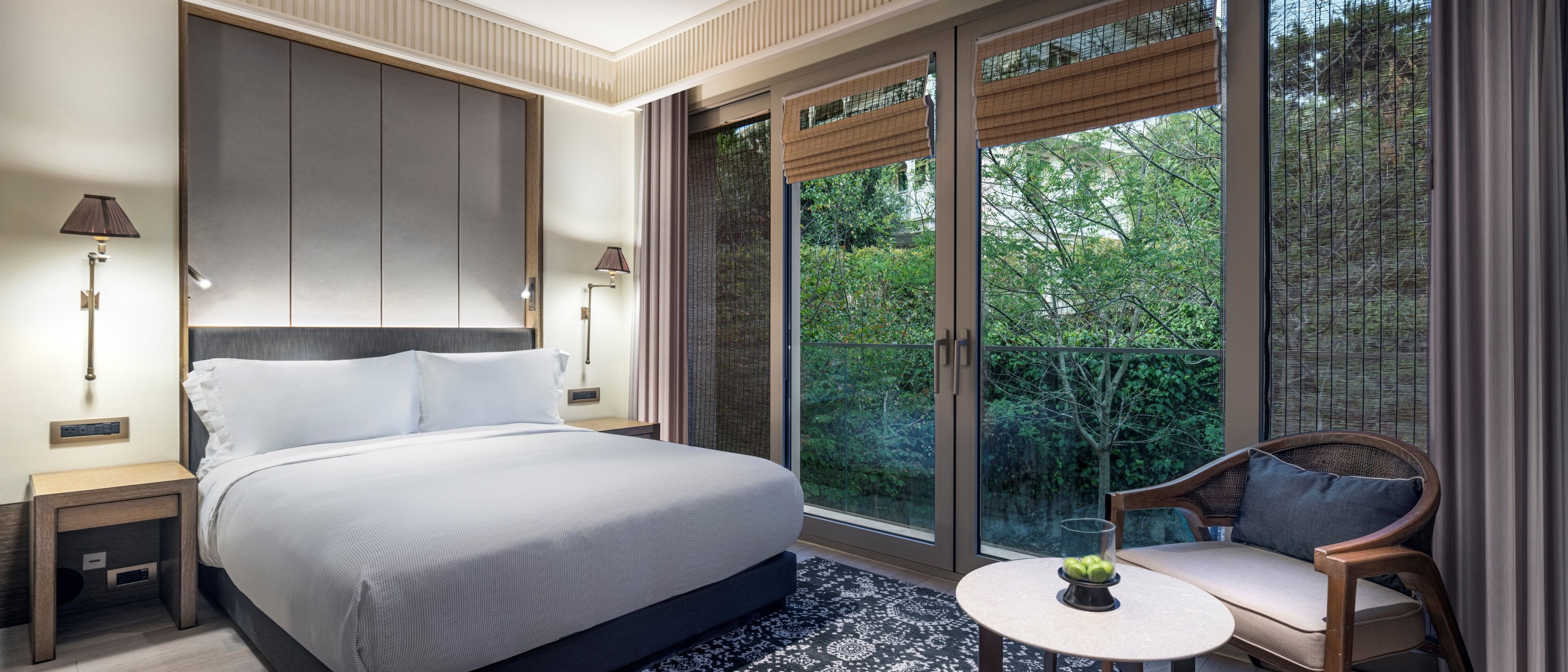 Un dormitorio con una cama grande, lámparas de noche, una mesita con frutero y vistas a zonas verdes a través de amplias ventanas.