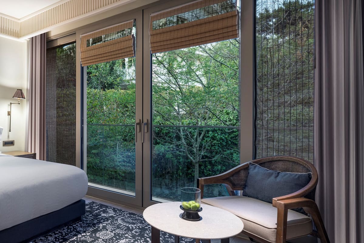 A bedroom with a large bed, bedside lamps, a small table with a fruit bowl, and a view of greenery through large windows.