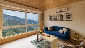 A room with a large window offering a mountain view, a blue sofa, a small table with a vase and books, and an air conditioning unit on the wall. - Soul Stories - Luxury Villas and Resort, Mussorrie (Dhanaulti)