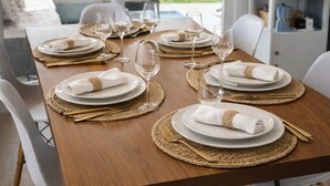 Un tavolo da pranzo apparecchiato con piatti bianchi, sottopiatti dorati e bicchieri di vetro trasparenti, illuminato da lampadari in vimini.