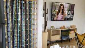 Un tavolo da pranzo apparecchiato con piatti bianchi, posate dorate e sottopiatti intrecciati. A parete, un televisore mostra la serie 'EMILY IN PARIS'.