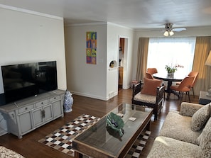 Een woonkamer met een flatscreen televisie, een glazen salontafel, een bank en een eethoek met een tafel en stoelen.