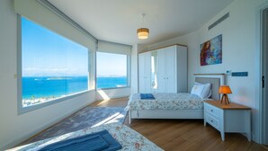 A bedroom with a large window offering a view of the sea, a bed with a floral bedspread, a bedside table with a lamp, and a wardrobe. - Villa Valentino 1 (Bodrum)