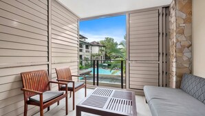 A balcony with a sofa, two wooden chairs, and a table. A pool and buildings are visible in the background.