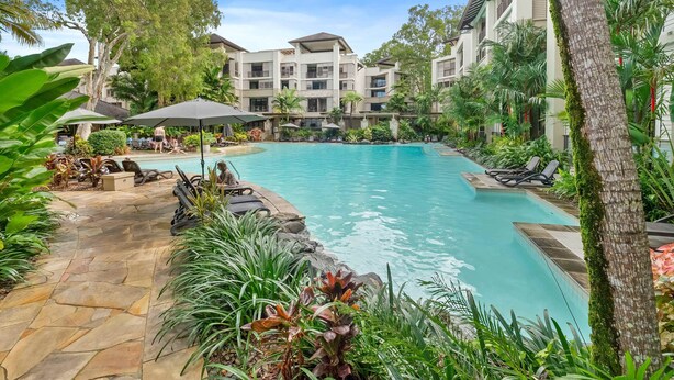 A pool area with lounge chairs and umbrellas, surrounded by lush greenery and a multi-story building in the background.