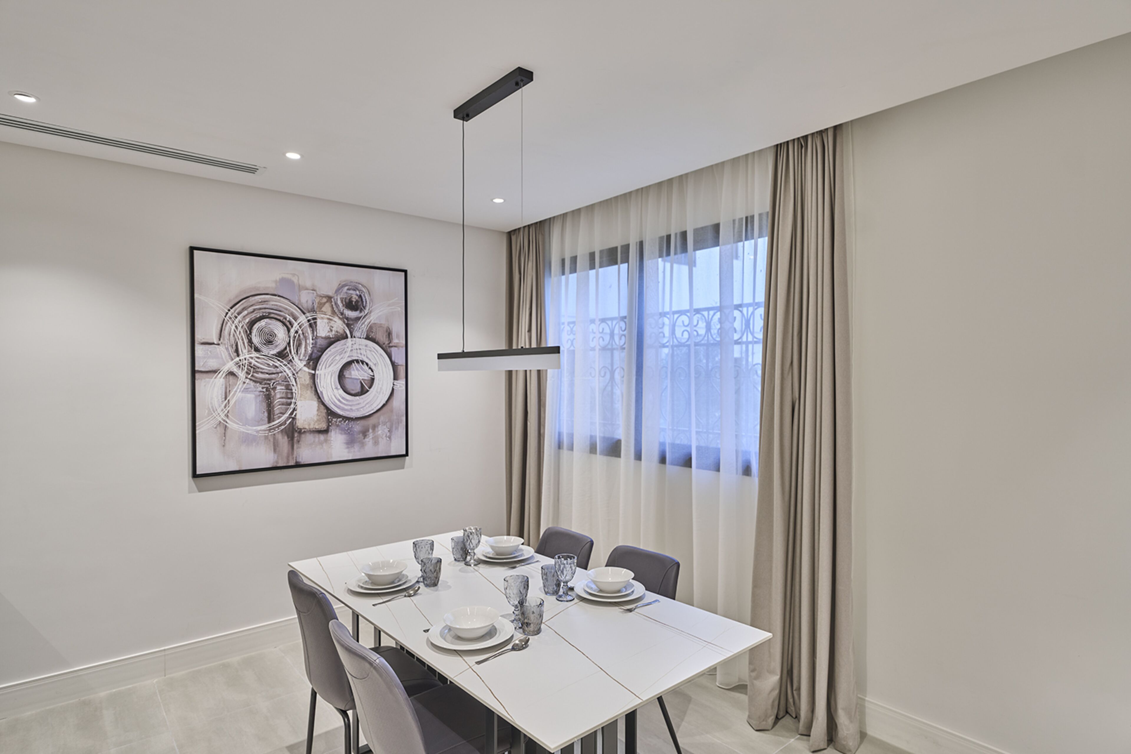 A modern dining room with a white table set for four, a contemporary painting on the wall, and a large window with curtains.