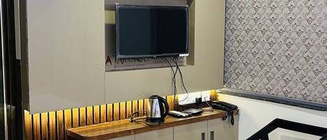 A modern hotel room with a large flat-screen TV mounted on a wall, a wooden bench with built-in storage, a black chair, and a marble-patterned floor.