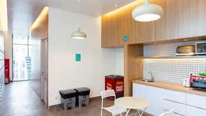 Eine moderne Küche mit Holzschränken, einer gefliesten Spritzschutzwand und einer Mikrowelle. Zwei Mülleimer und ein Tisch mit Stühlen sind ebenfalls vorhanden.