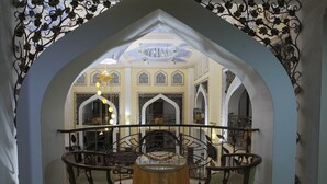 A dining area with a round table and chairs, ornate metal railings, and a view of a room with a chandelier and patterned ceiling.