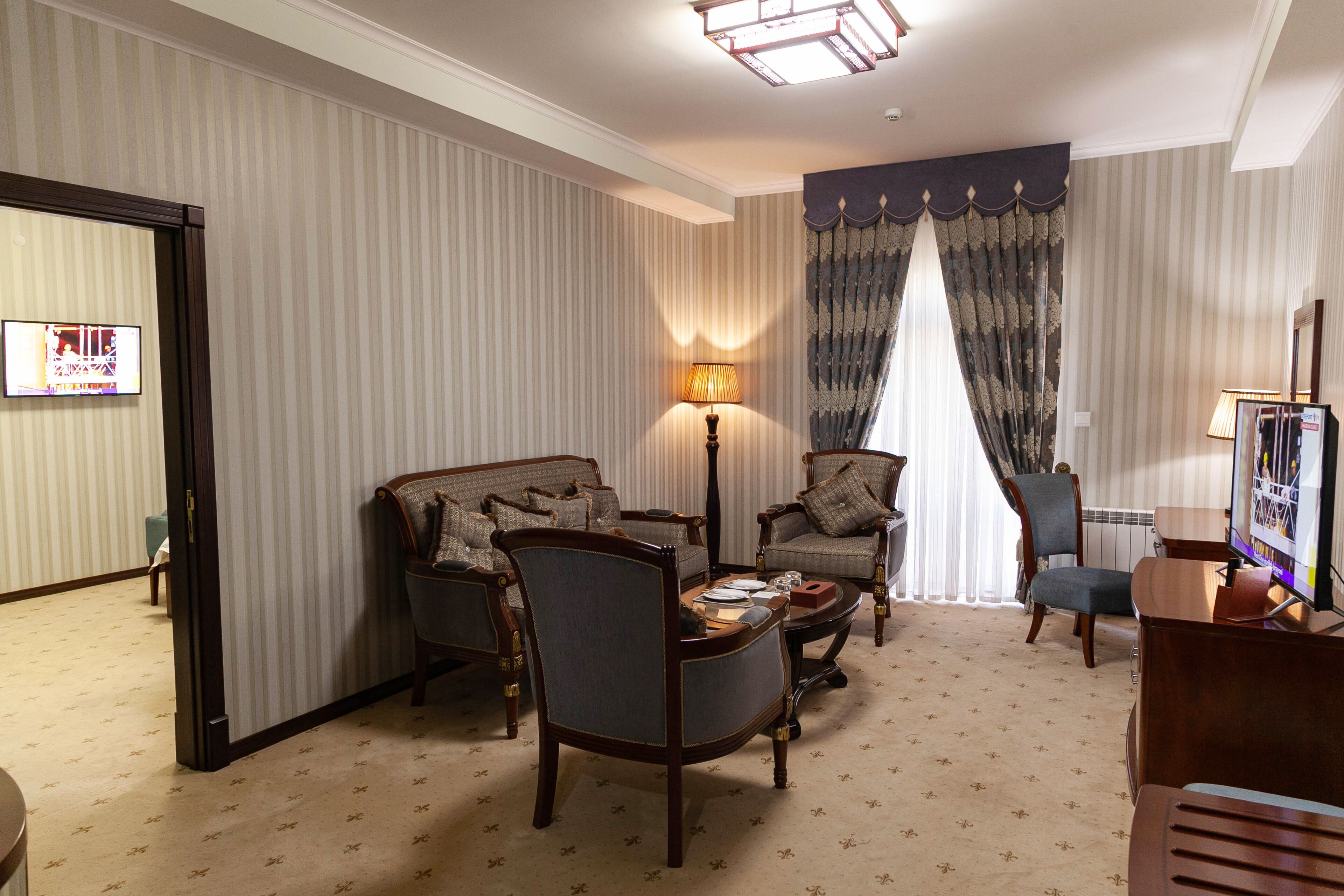 A hotel room with striped wallpaper, a large window with curtains, a flat-screen TV, and a seating area with two armchairs and a sofa.
