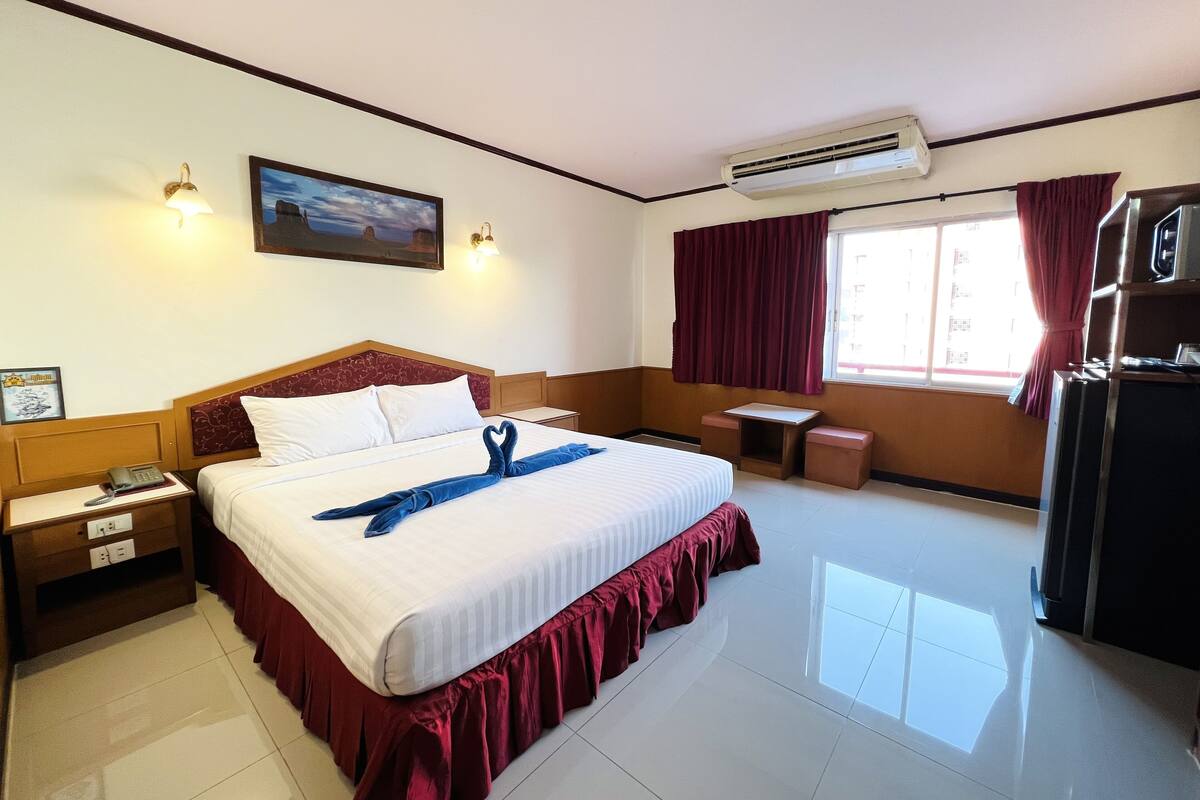 A hotel room with a large bed, a bedside table with a phone, a small table, an air conditioning unit, and a window with curtains.