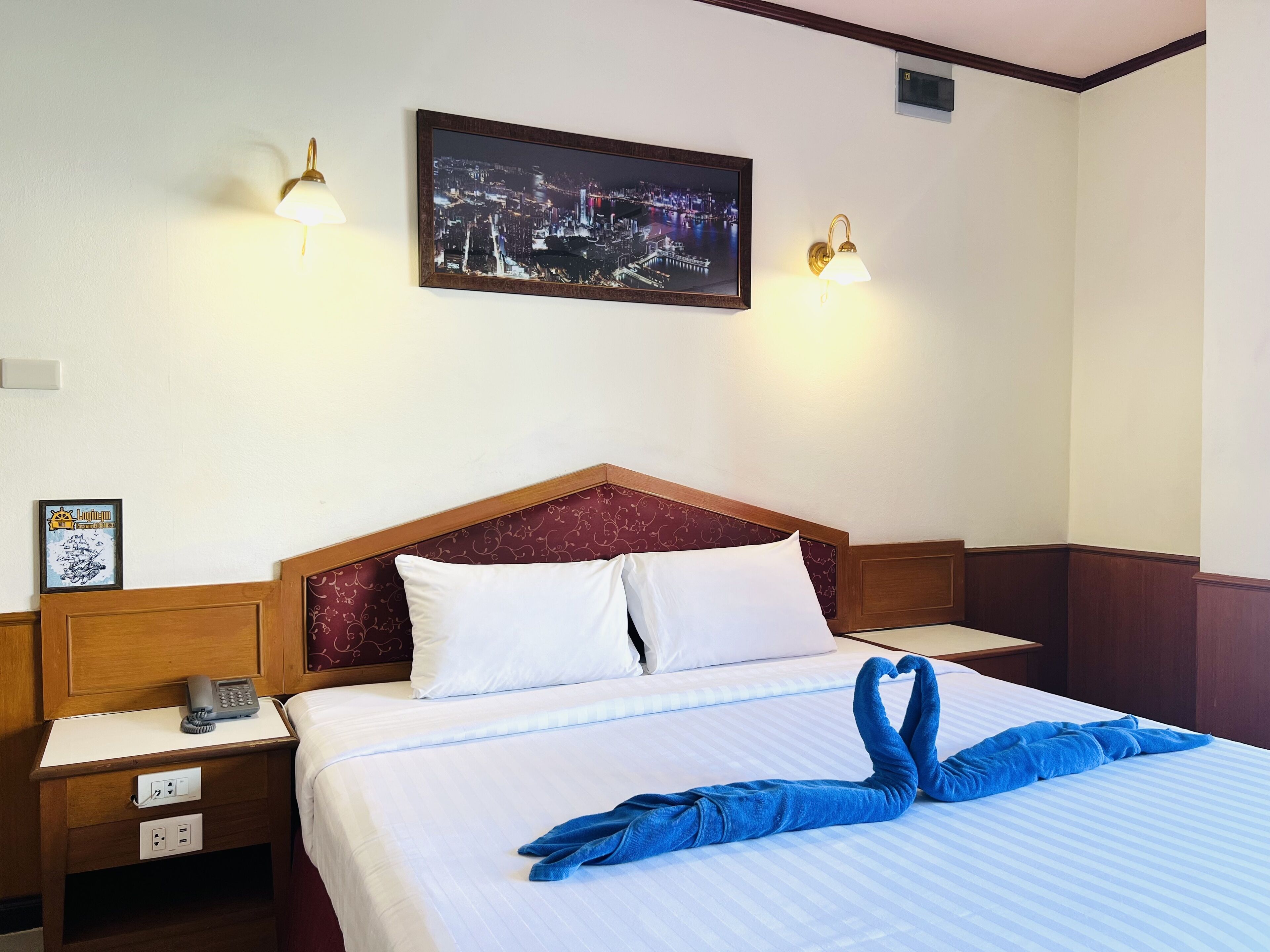 A hotel room with a bed, a nightstand with a phone, two wall-mounted lamps, and a framed cityscape picture.