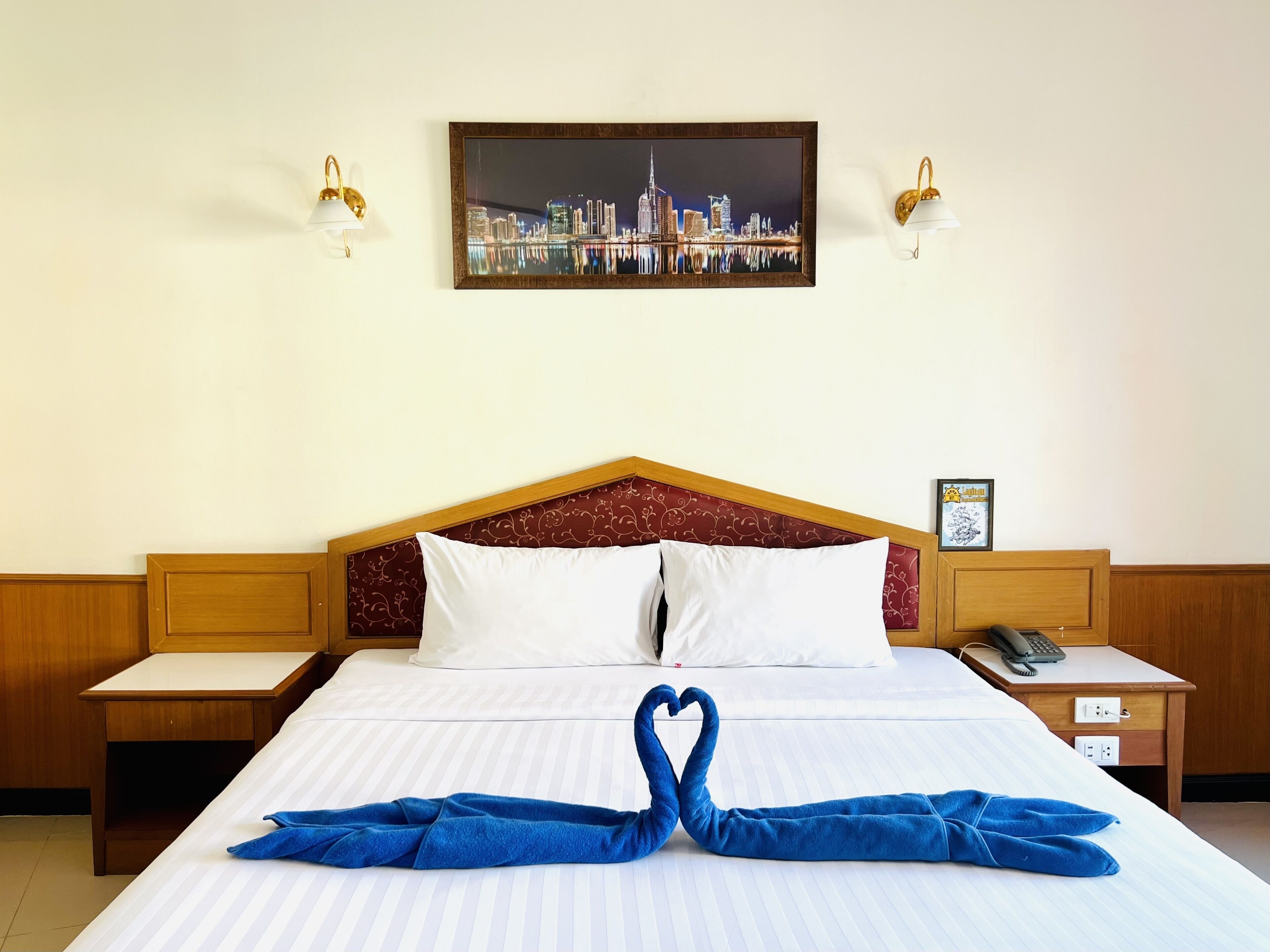 A hotel room with a bed, two bedside tables, a wall-mounted picture, and a heart-shaped towel decoration.