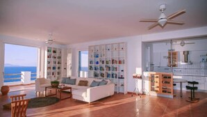 A spacious living room with a white sofa, a coffee table, and a bookshelf filled with books. There is a view of the sea through large windows.