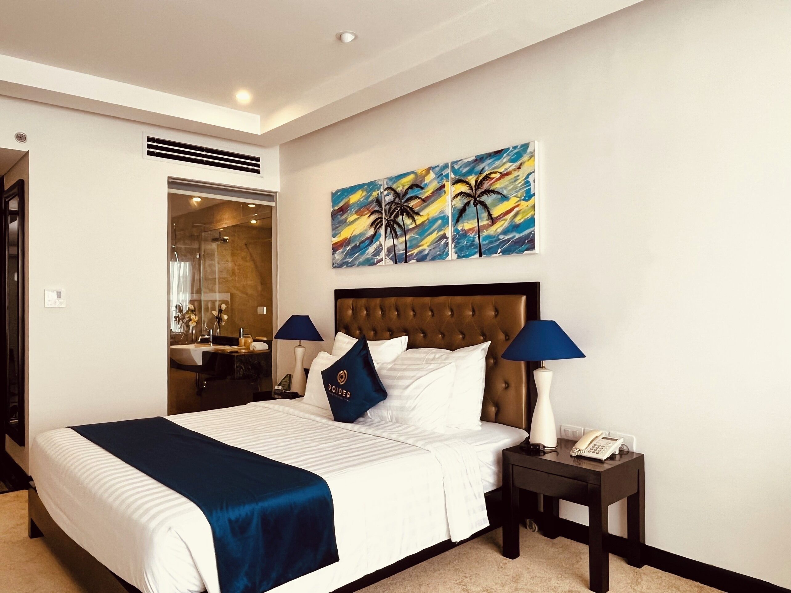 Una habitación de hotel con una cama grande, dos lámparas de noche, una mesita de noche con un teléfono y un cuadro de palmeras en la pared.