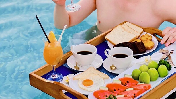 A person in a swimming pool holding a drink, with a tray containing food and beverages.