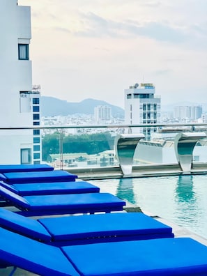 A rooftop pool area with blue lounge chairs, a view of the cityscape, and a bridge in the background. - Sandals Island Hotel  (Nha Trang)