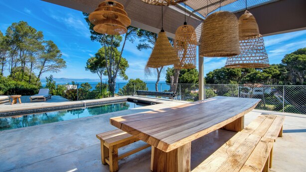 Una mesa de madera junto a la piscina con bancos, rodeada por una alberca y con vistas a un paisaje pintoresco de árboles y el mar.