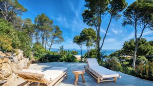 Terraza de madera con dos sillas reclinables y una pequeña mesa, con vista a un paisaje costero con árboles y el mar.