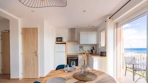 Une cuisine moderne avec des meubles blancs, une table à manger en bois surmontée d’un set de table en fibres tressées, et une vue sur la plage grâce à de grandes fenêtres.