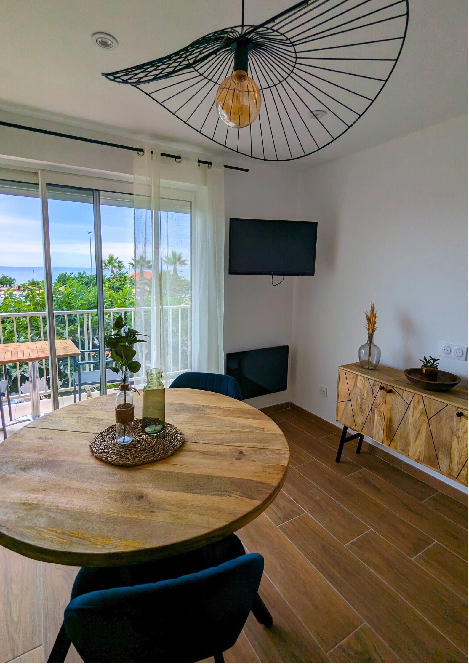A modern room with a round wooden table, a TV mounted on the wall, and a balcony with a view of the sea.