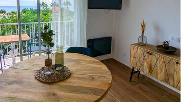 A modern room with a round wooden table, a TV mounted on the wall, and a balcony with a view of the sea.