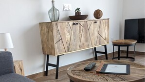 Un salon moderne avec un canapé gris, une table basse ronde en bois sur laquelle se trouvent une télécommande et un carnet, un meuble en bois agrémenté d’objets décoratifs et un meuble TV noir.