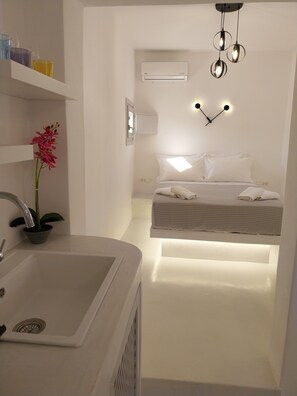 A modern, minimalist bedroom with a built-in bed, a bedside table, a wall-mounted air conditioner, and a pendant light fixture. - East Wind Perissa (Santorini)