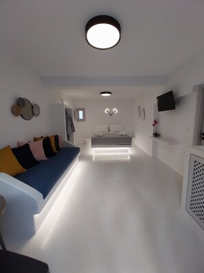 A modern, well-lit living area with a sofa, a bed, and a TV mounted on the wall. - East Wind Perissa (Santorini)