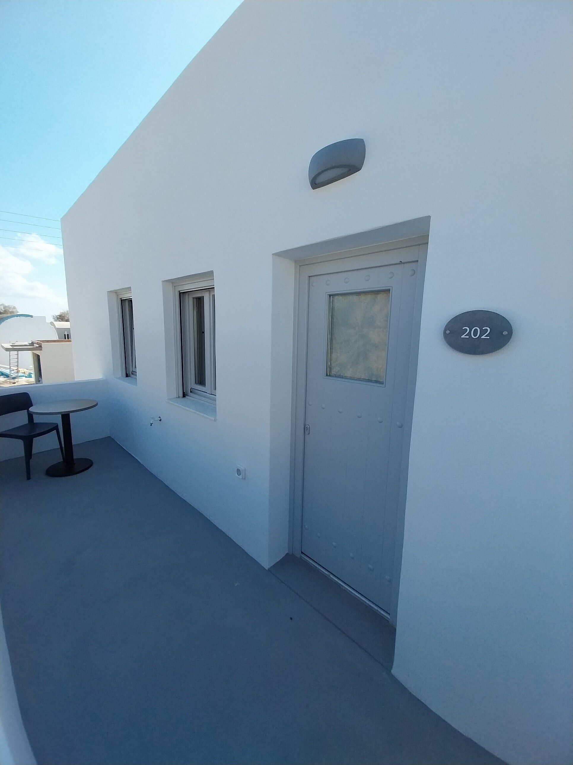 A white building with a door marked '202', a small window, and a table with a chair.