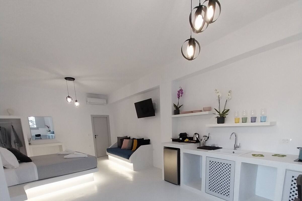 Ein moderner, minimalistischer Wohnraum mit einem großen Bett, einer kleinen Sitzecke, einer Küche mit Spüle und einem an der Wand montierten Fernseher.