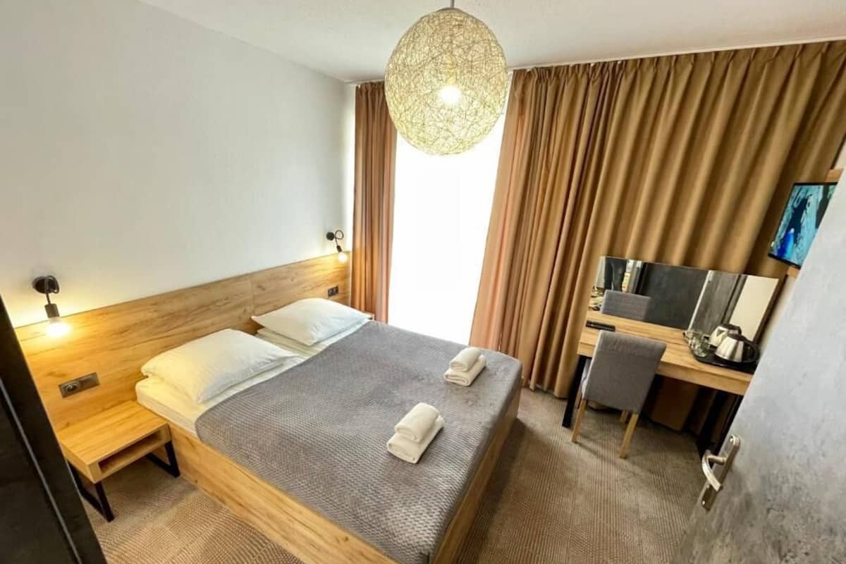 Quarto de hotel com cama de madeira, uma escrivaninha, uma cadeira, televisão e uma janela grande com cortinas.