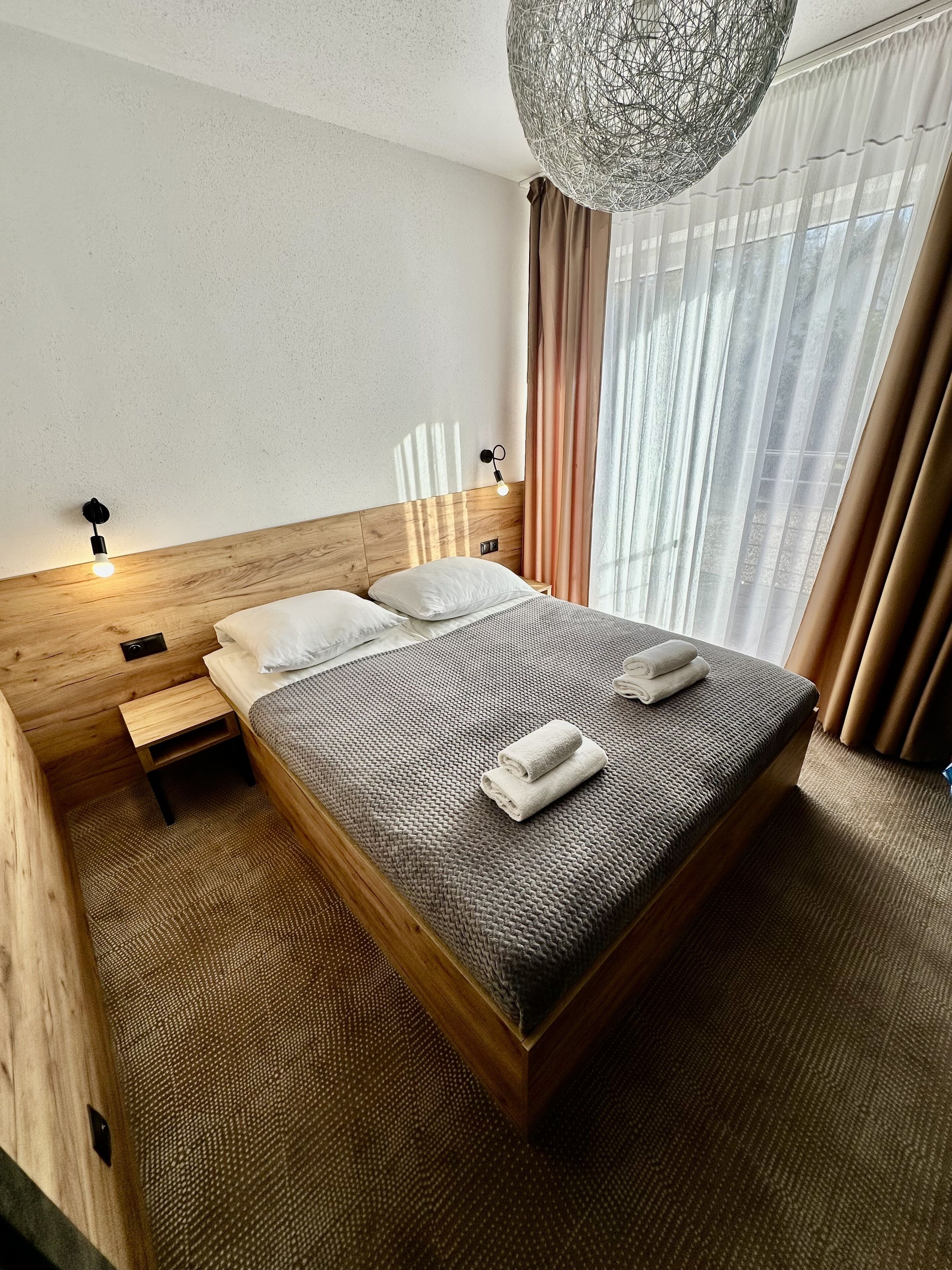 A hotel room with a wooden headboard, a large bed with a textured gray bedspread, two white pillows, a bedside table with a lamp, and a hanging light fixture.