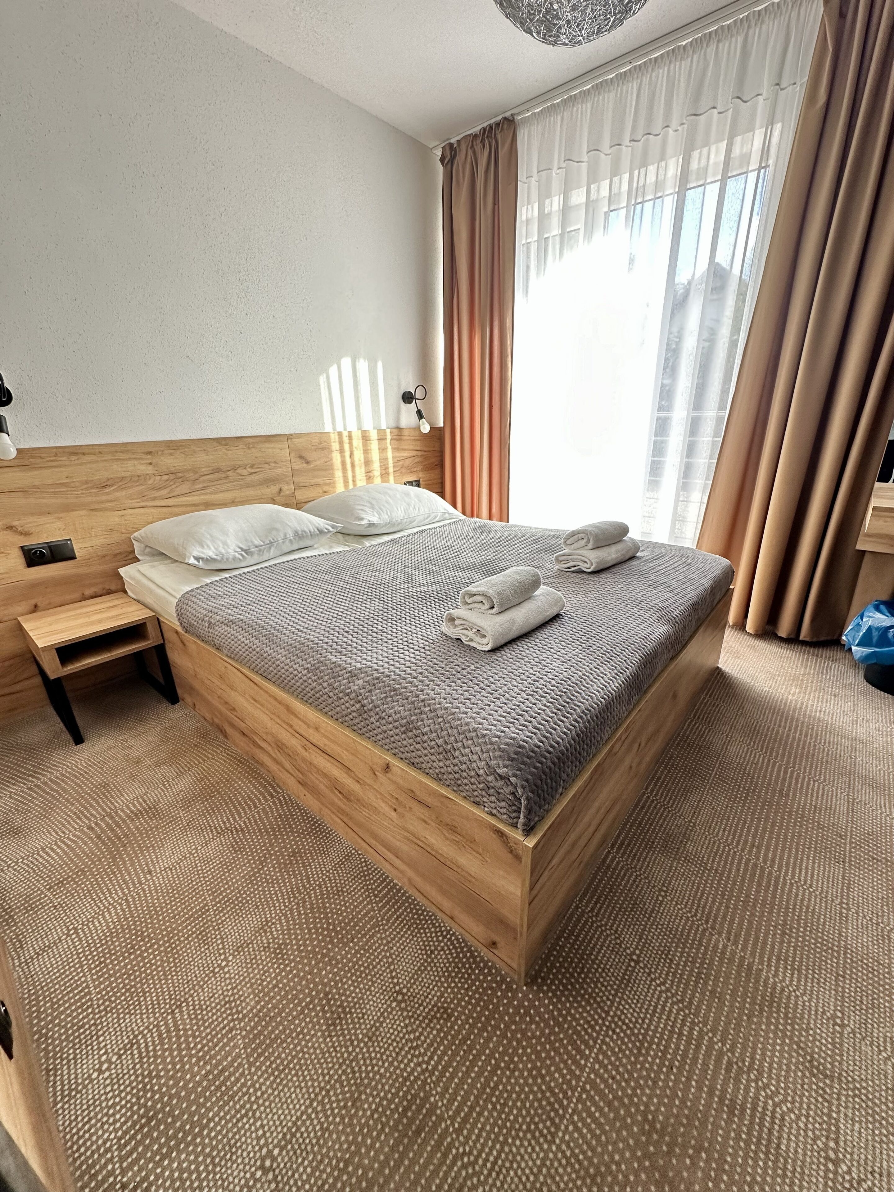 A neatly made bed with a textured blanket and folded towels, a wooden headboard, a bedside table with a lamp, and a view of the outdoors through sheer curtains.