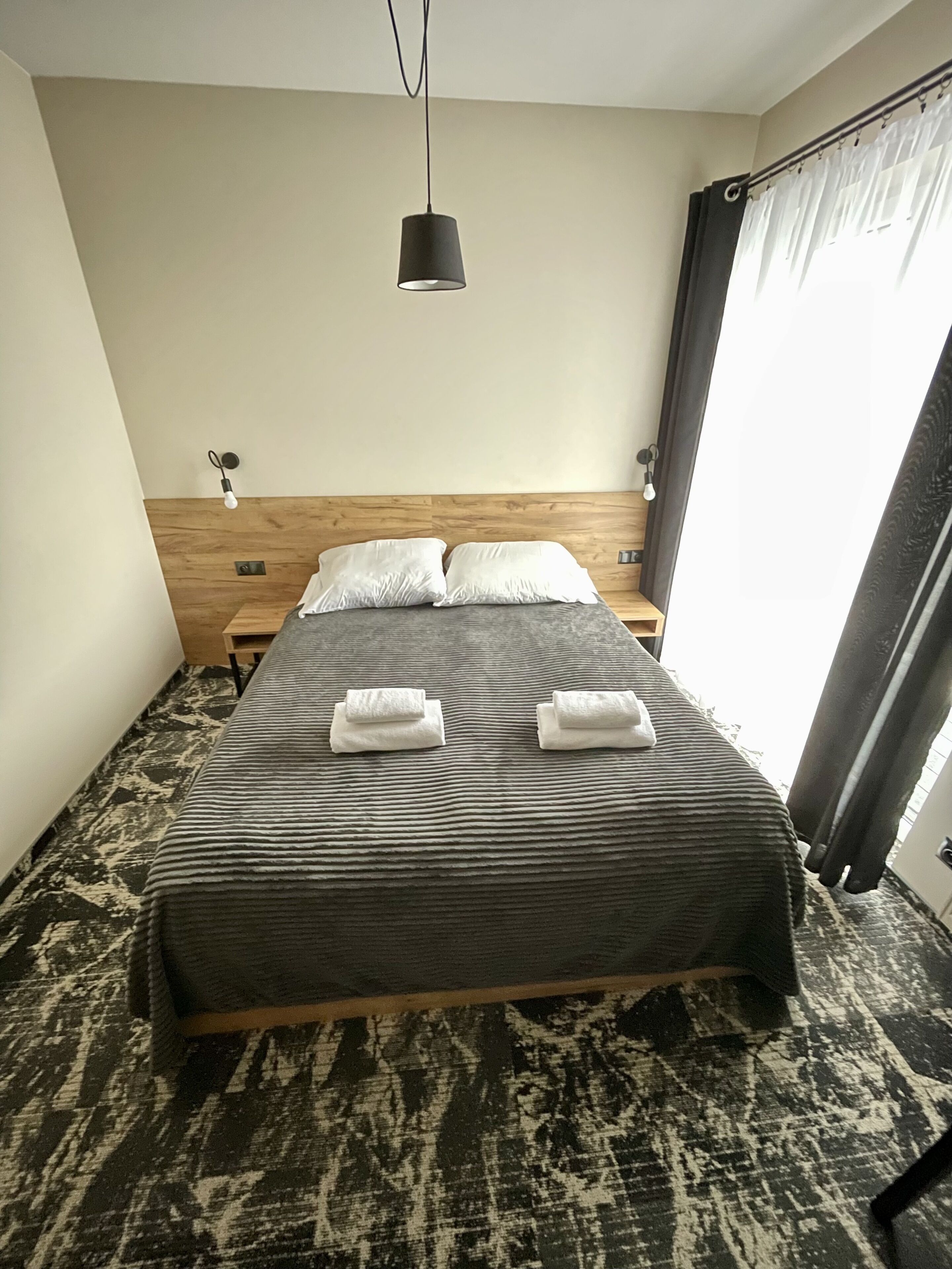 A neatly made bed with a dark patterned bedspread and two pillows, a wooden headboard, bedside tables with lamps, and a large window with curtains.