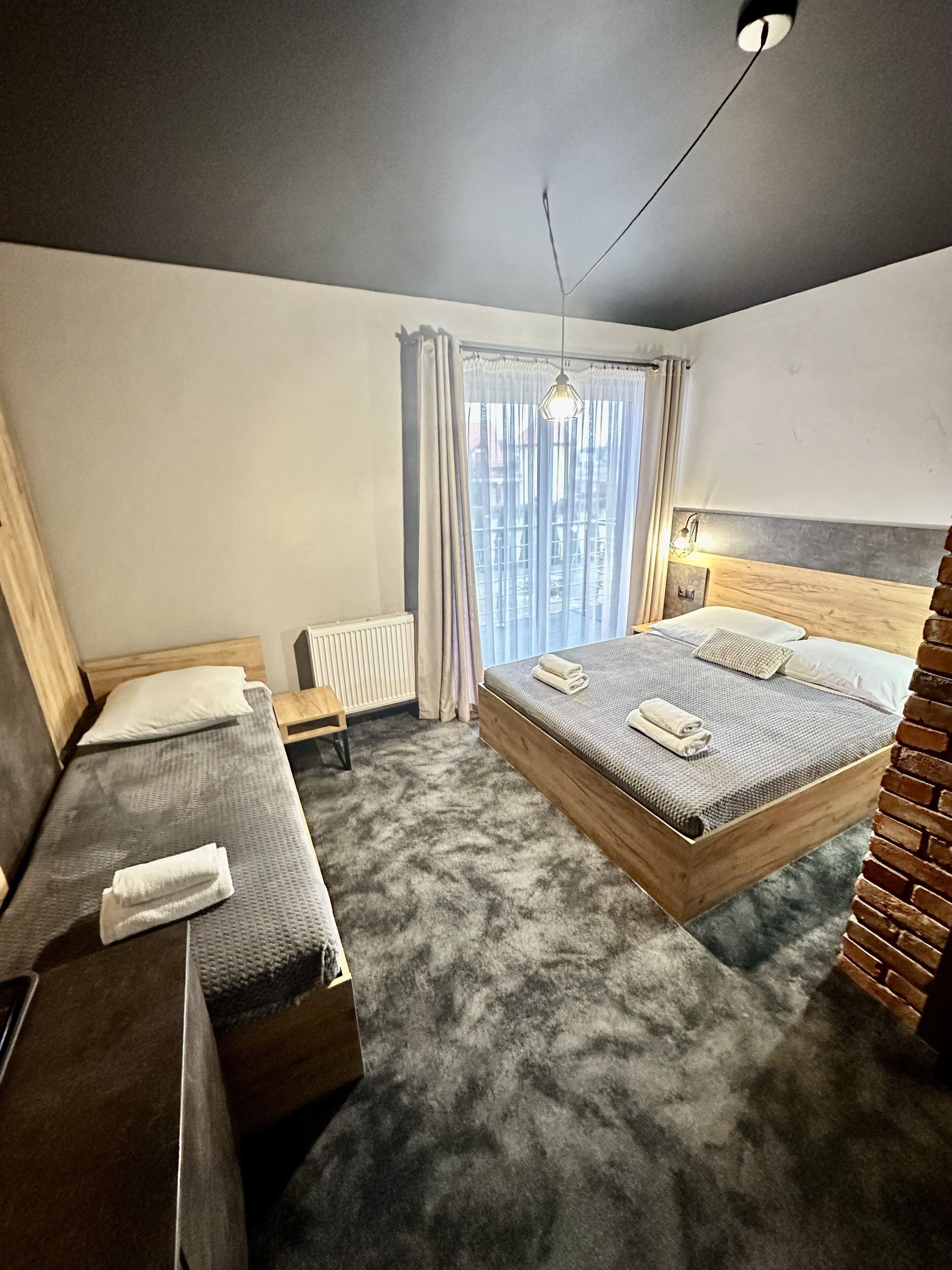 Et moderne hotelværelse med en stor seng, en mindre sofa, en træsenggavl, en radiator og udsigt over byen gennem lette gardiner.