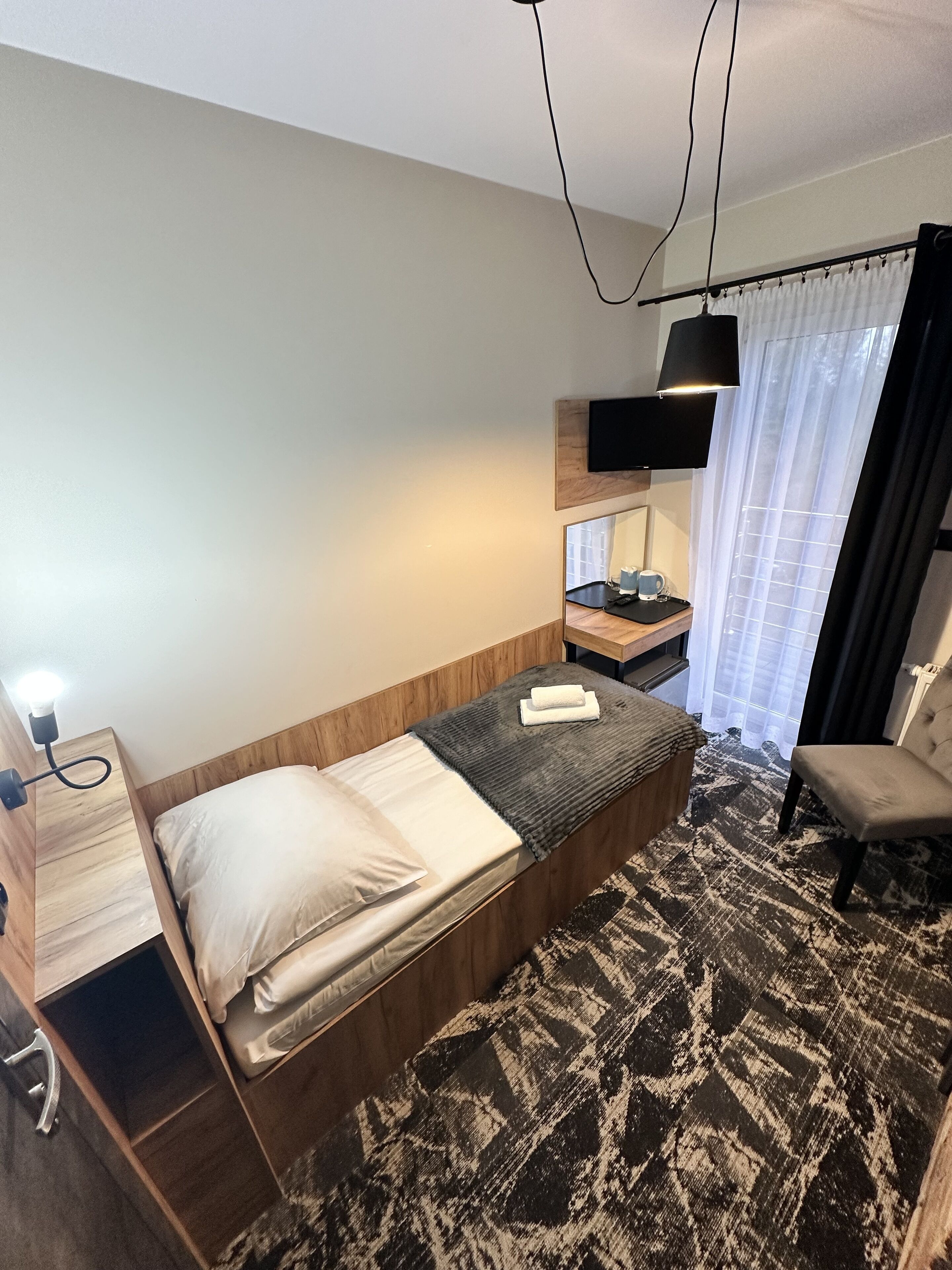 Quarto de hotel com cama de madeira, mesa de cabeceira com luminária, cadeira, televisão fixada na parede e janela com cortinas.