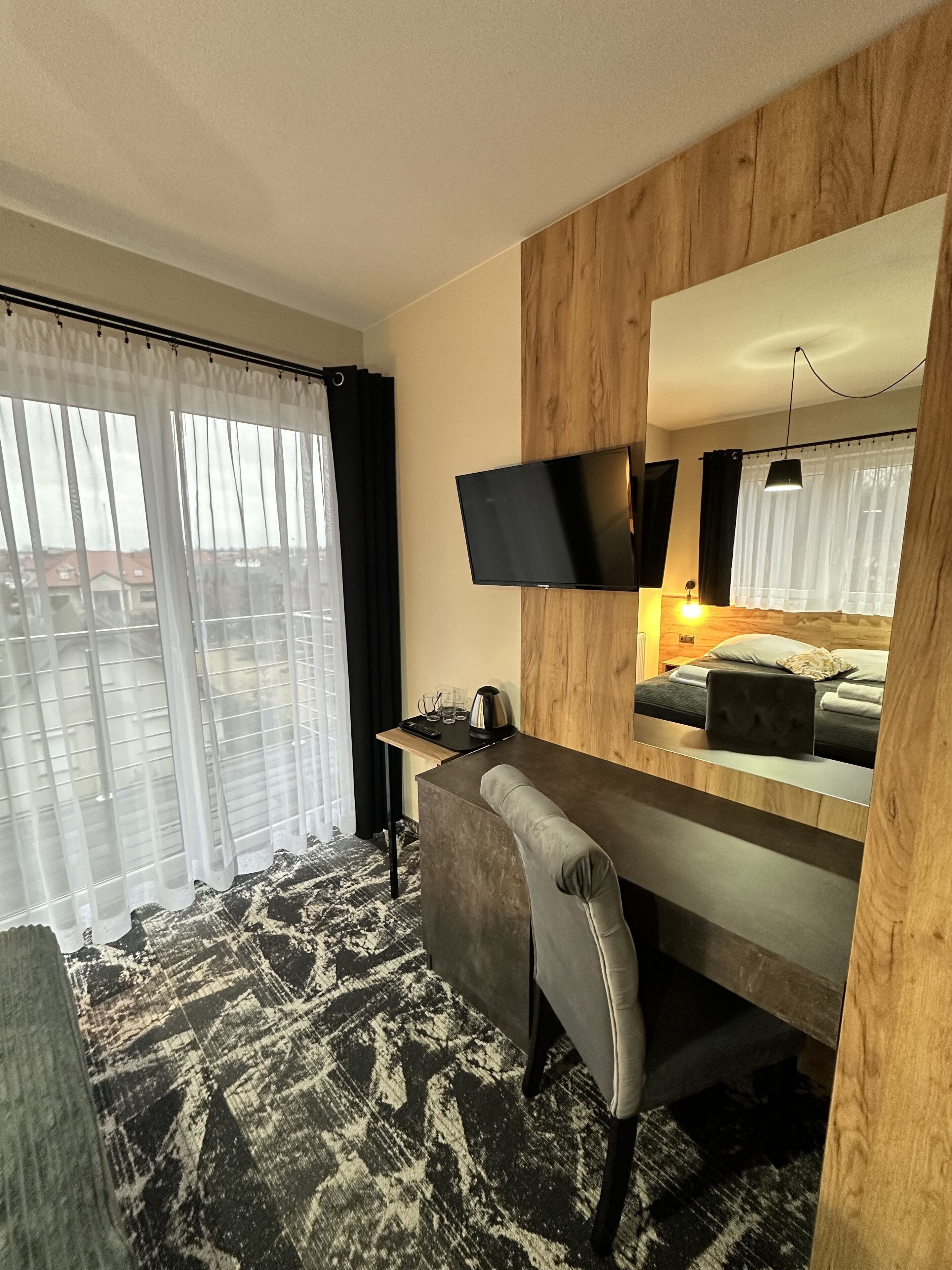 Una camera d'albergo con un letto, una scrivania, una sedia, una TV fissata al muro e un'ampia finestra con tende leggere.